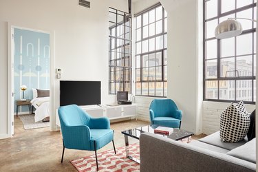 Modern loft living room with gray couch, blue accent chairs, white console, mirror coffee table, large floor lamp, industrial windows, orange pattern rug.